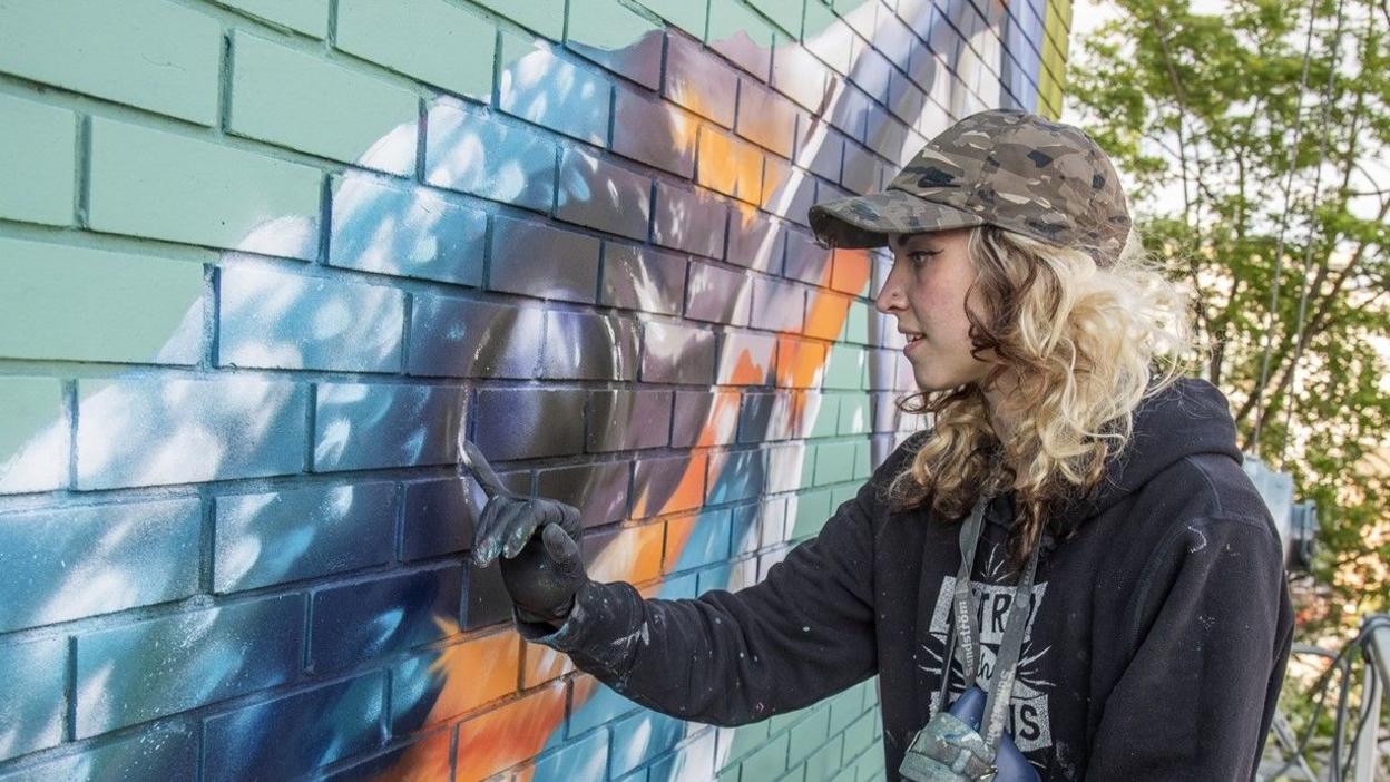 A close-up of artist Megan Russell, working on her nominated spray painting, "Reverie". From here we can just see part of the kingfisher's head, she is touching its eye to add white paint, on a brick wall. It has light blue, dark blue and orange colours on its face. There are trees in the background showing she must be high up. Megan is in her early thirties. She has mostly bleached blonde hair with some brown streaks at the front and underneath. Her hair is curly. She is wearing a camouflage-printed hat and a black hoodie.