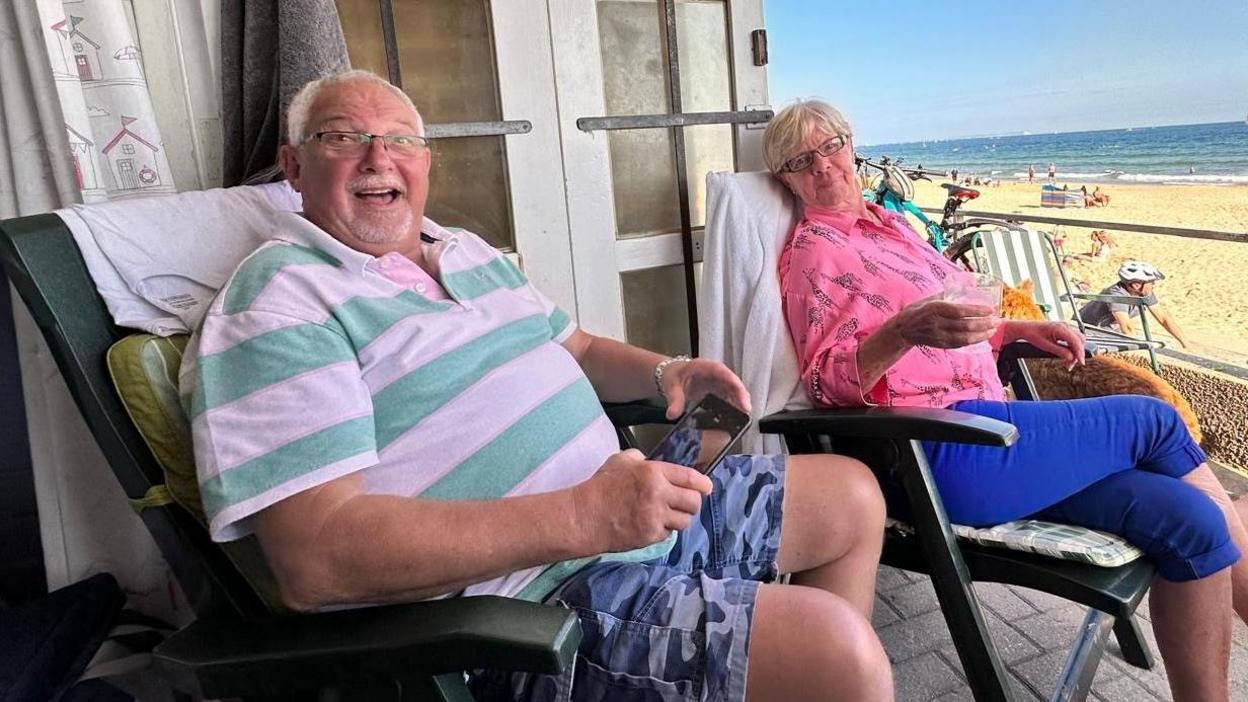 A man and a woman in their seventies sitting on chairs outside a beach hut. The woman is wearing a pink top with blue cropped trousers and is holding a drink. The man is grinning and wearing a blue and white-striped top with blue camouflage-print shorts. In the background is a sandy beach with blue sea. It looks sunny.