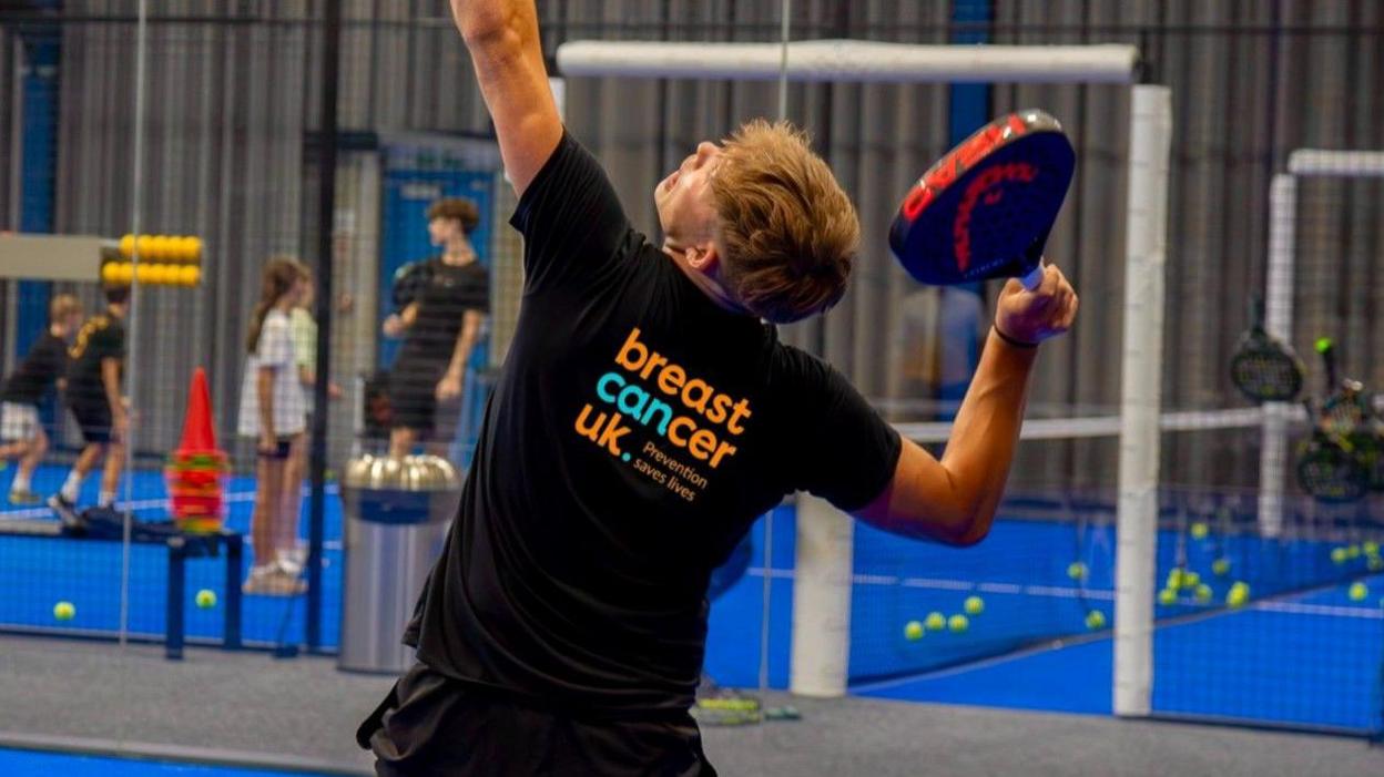 A man playing Padel on a court. He is wearing a black T-shirt and shorts with the words Breast Cancer UK written on the back.