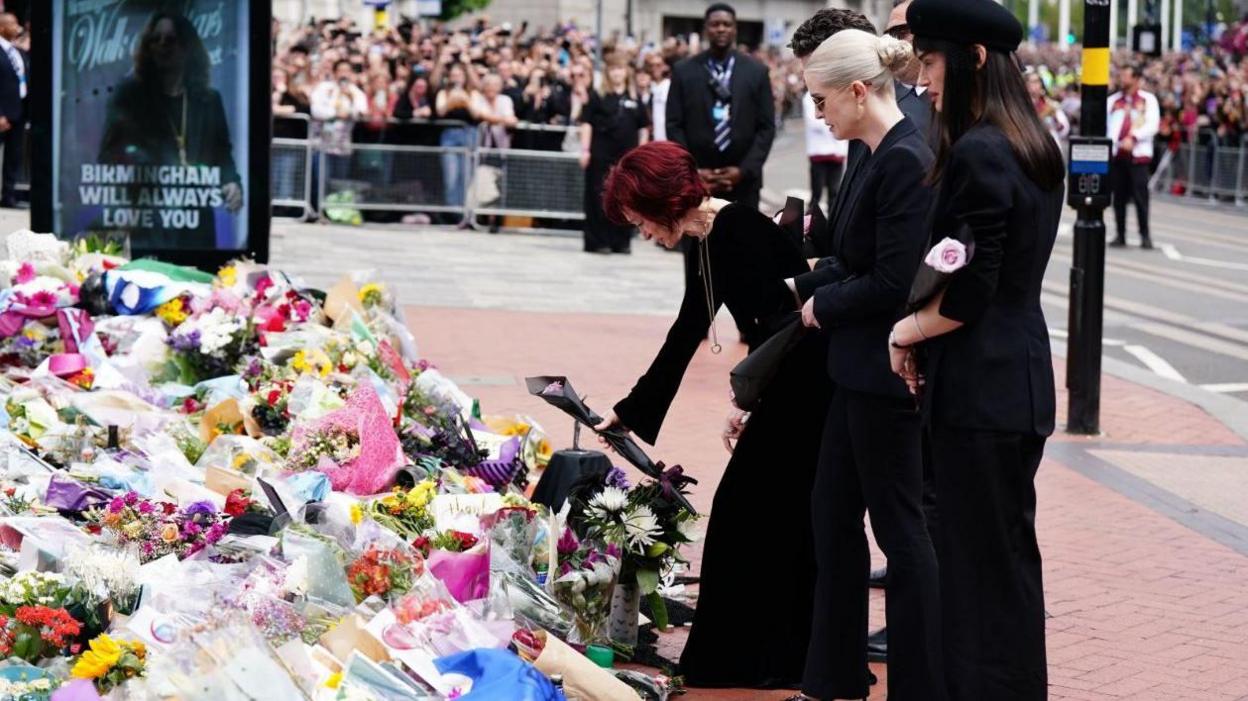 There are dozens of flowers, wrapped in cellophane, at the left hand side of the picture with a poster of Osvbourne behind with the words Birmingham will always love you. A woman dressed in black with dark red heair, lays a pink rose wrapped in black paper down on them. She is with three other people also in black.