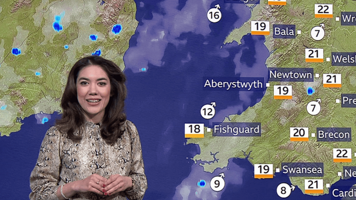 A brown-haired woman reporting on the weather, stood in front of a green screen displaying the map of Wales. She is wearing a snake print blouse.