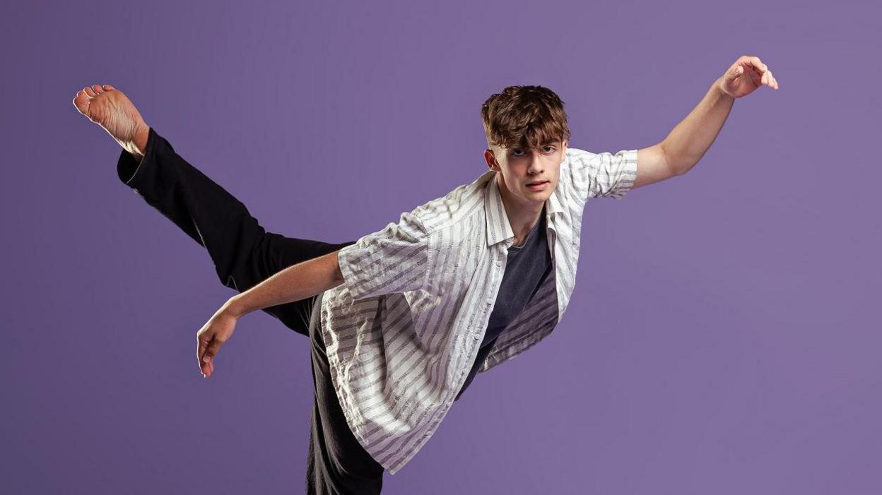Joe is performing a dynamic dance pose against a solid purple background. The individual is balancing on one leg with the other leg extended high behind them, arms outstretched for balance. They are wearing a loose, light-coloured striped shirt over a dark top and black pants.