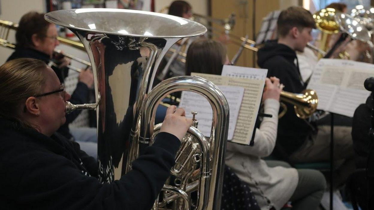 Members of Durham Miners’ Association band playing various instruments. They have sheets of music in front of them. A blonde woman with black-rimmed glasses is in the foreground, playing a silver tuba.