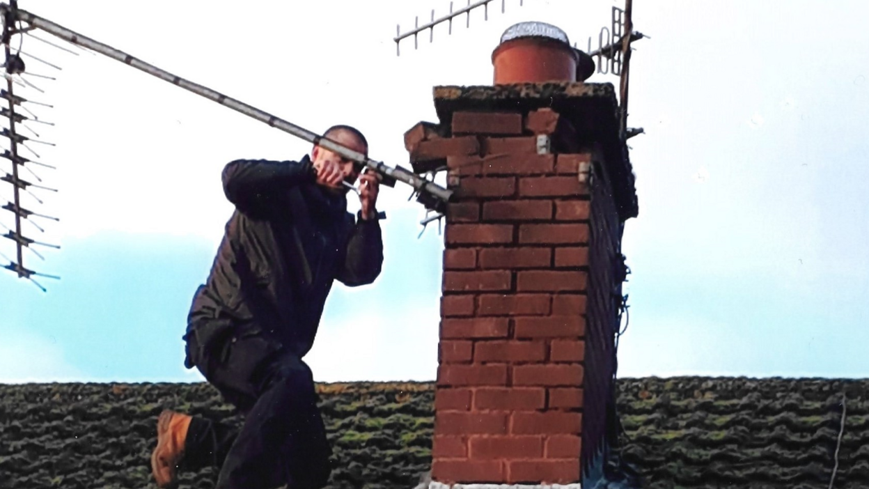 A man in black clothing and brown boots kneels on a tile roof, working on an aerial which is attached to a chimney.