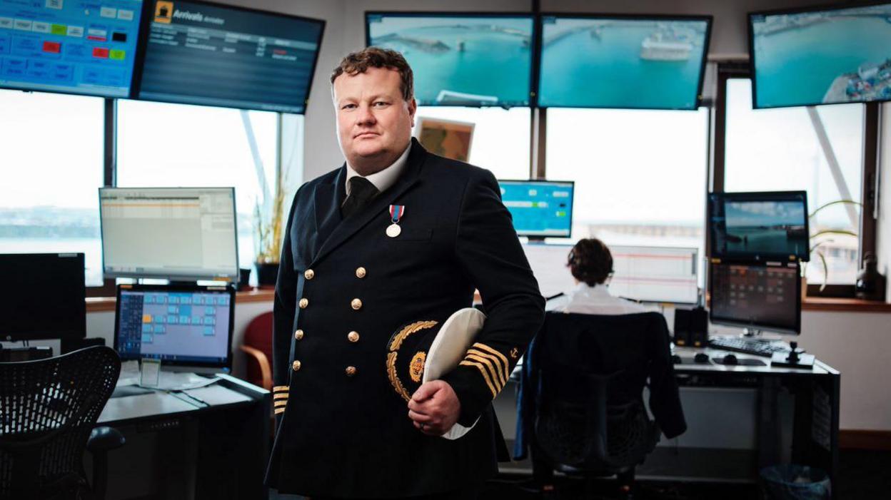 Captain Bill Sadler is wearing naval uniform with a medal and holding his hat under his arm in a port operations room which has many screens of text and graphics