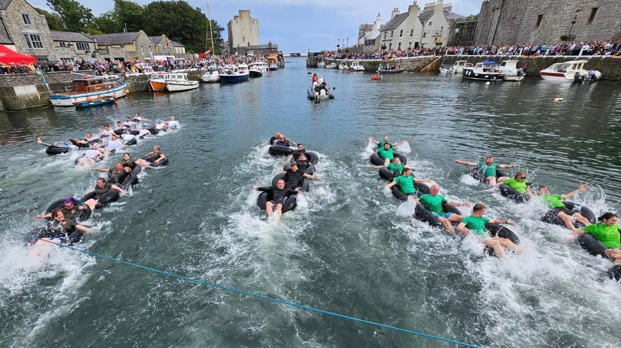 Teams of six in inflatable rings, which are linked together like a snake, racing in Castletown Harbour. The competitors are using their arms to paddle, which is creating lots of white splash. The harbour walls are lined with hundreds of spectators.