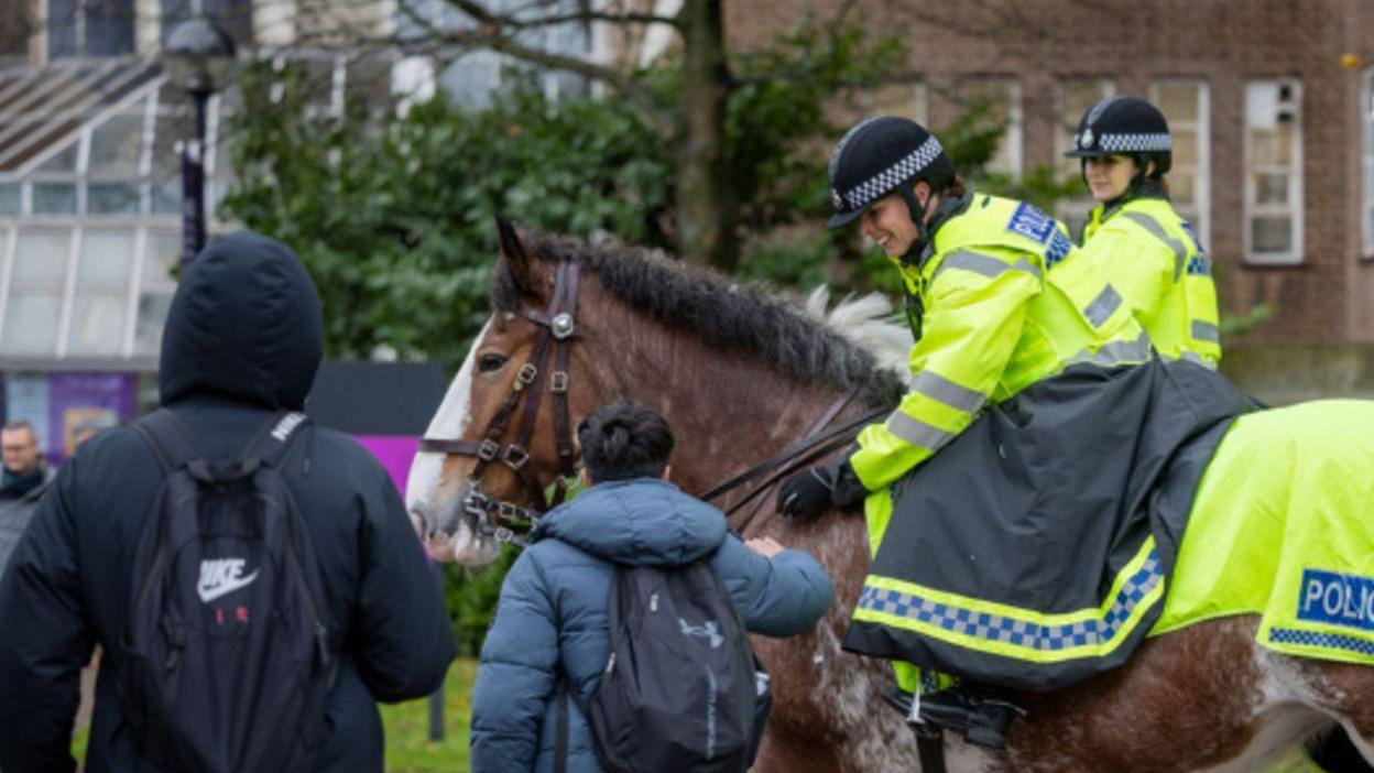 Two horses ridden by people in yellow reflective jackets and black riding helmets next to two people in dark coats