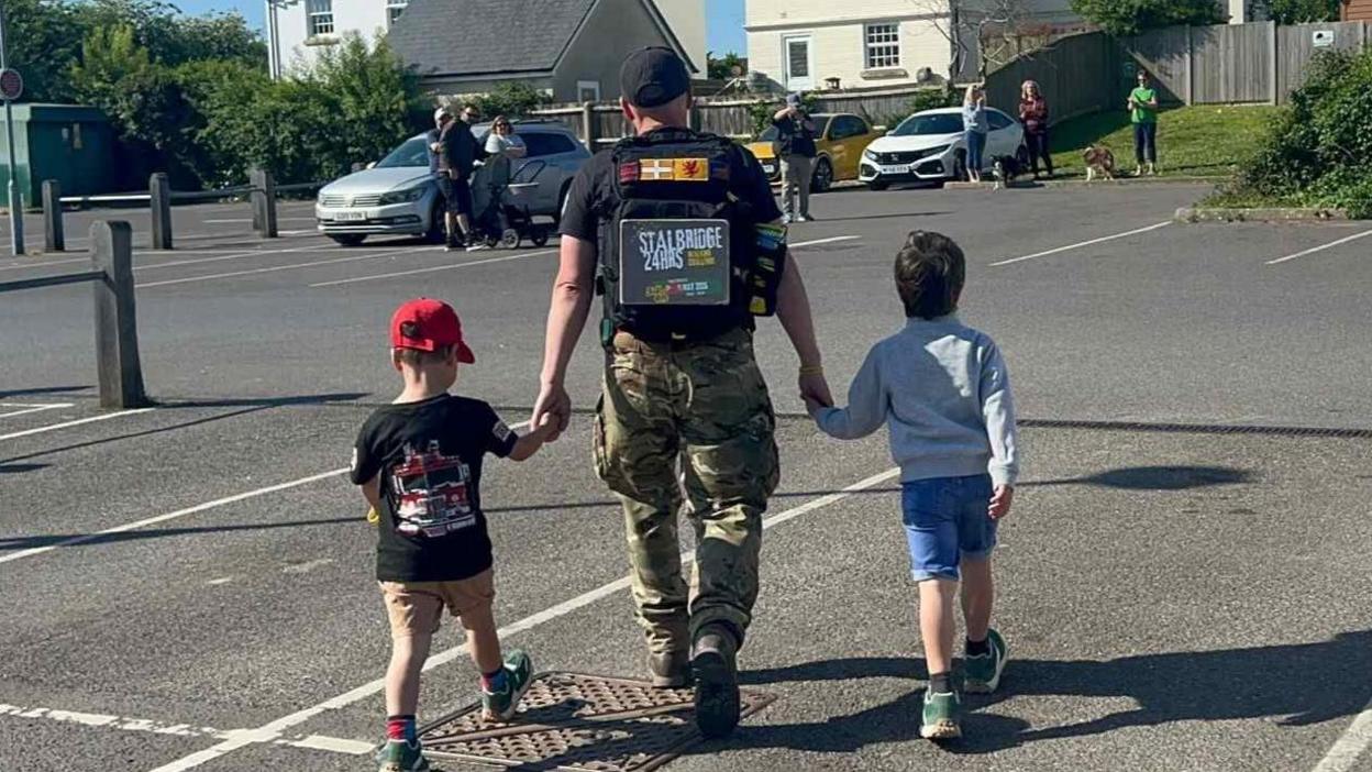 Martyn Harris has is back to the camera and his holding his two young sons hands. Martyn is wearing a black t-shirt advertising his Stalbridge 24 hours walking challenge and army trousers. His son on the left has a red cap on, black t-shirt and brown shorts while the one on the right has blue shorts and a white jumper.