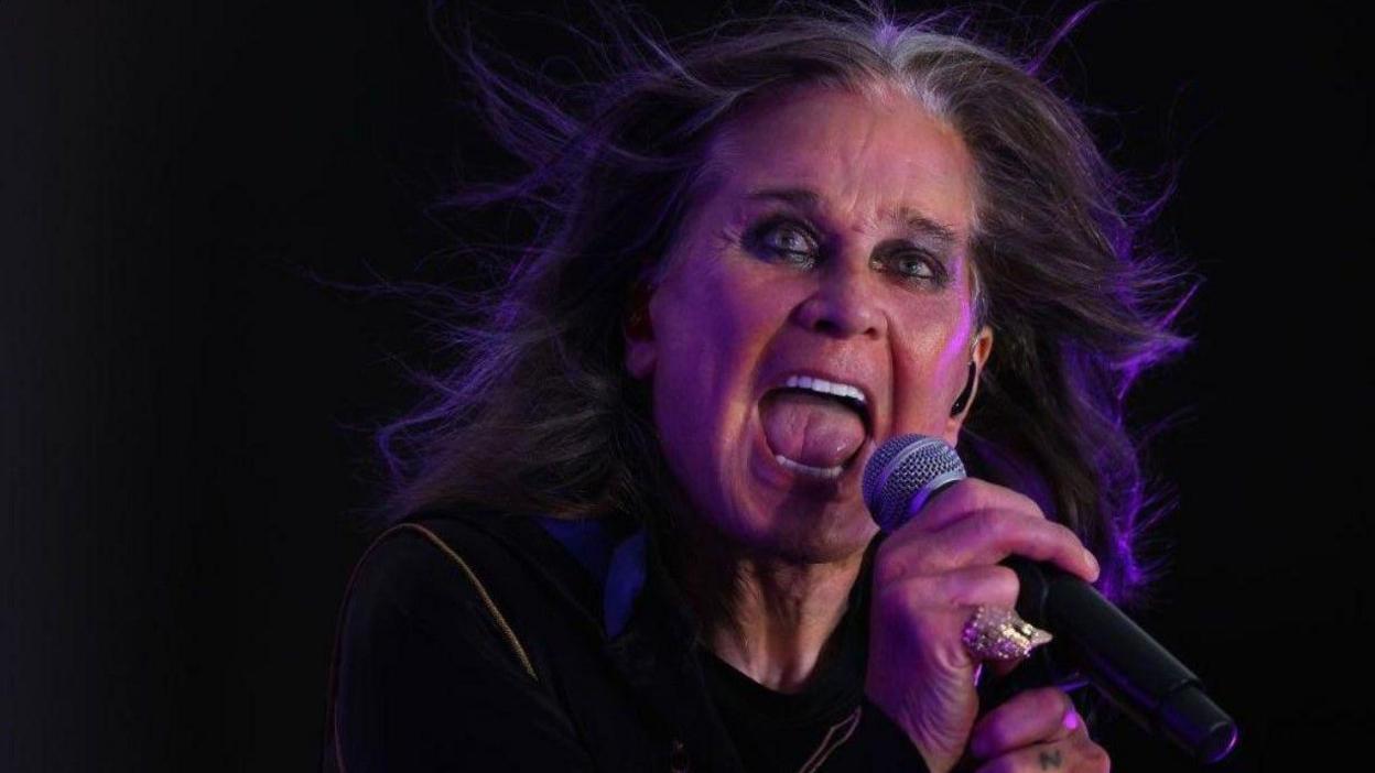 Ozzy Osbourne is holding a microphone with his right hand and has his mouth open. The background is black.