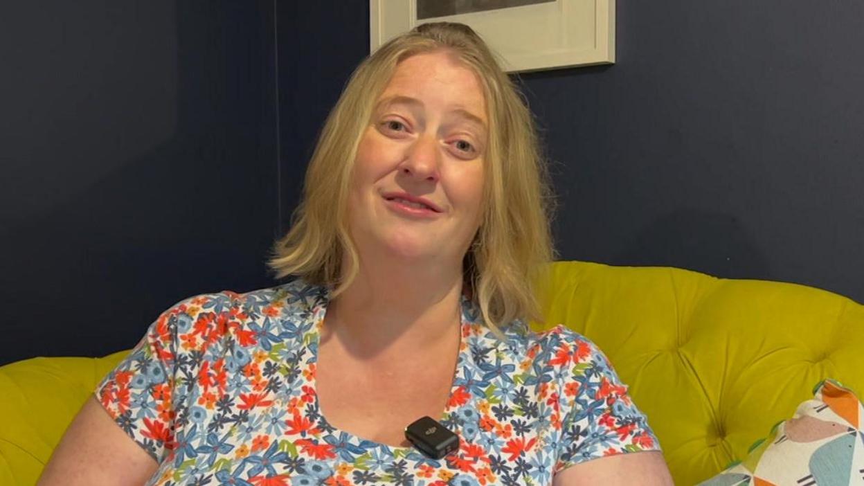 A lady with blonde hair wearing a floral multicoloured dress sat on a yellow sofa. She is looking straight at the camera sat in front of a blue wall with a white picture frame hung up behind her. 