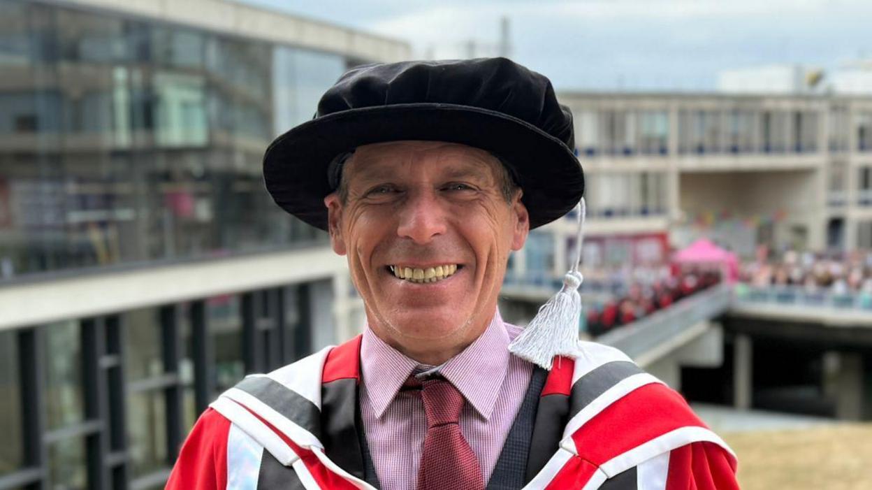 A smiling Peter Dutch looking straight at the camera. He is standing in front of a lecture hall wearing graduation robes over a dark grey suit, red shirt and red tie.