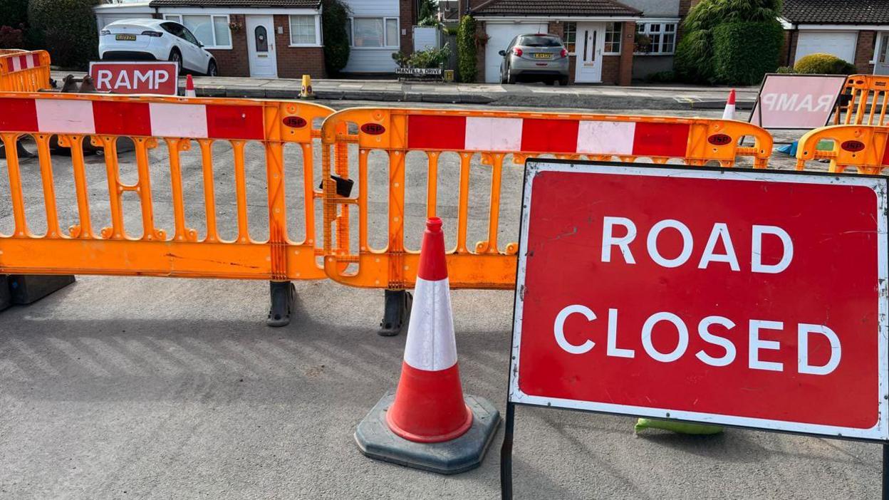 Orange barriers and cones at the side of road resurfacing works on a housing estate. A large red Road Closed sign is on the right.