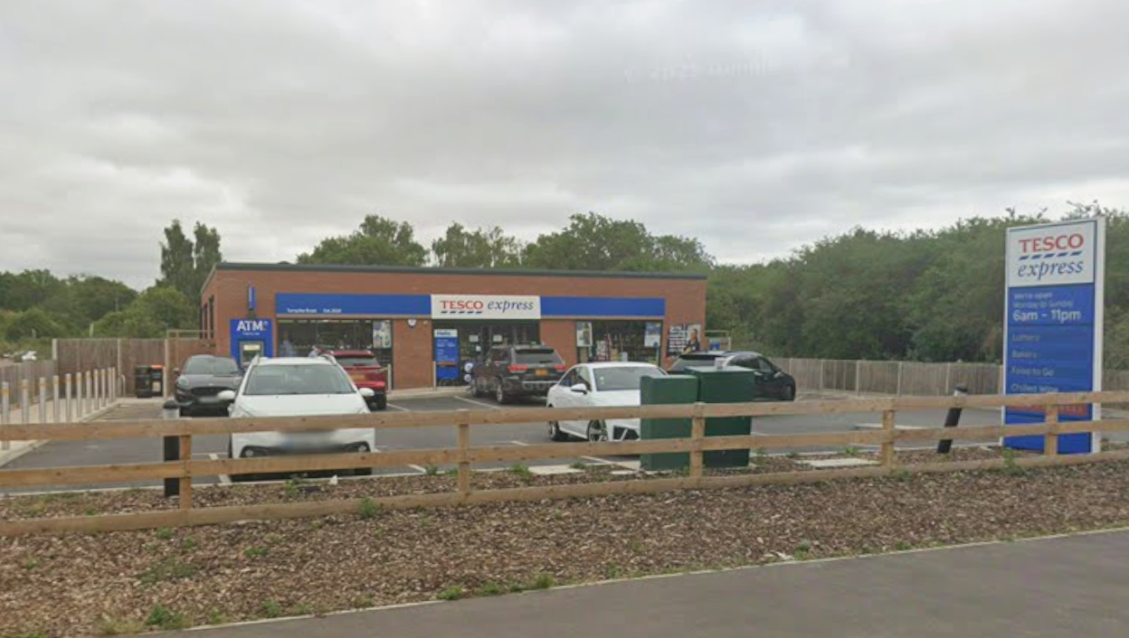 A small Tesco Express shop at the side of a road. A small car park sits in front of the shop with some vehicles parked up. A cash machine can be seen at the left hand side of the shop.