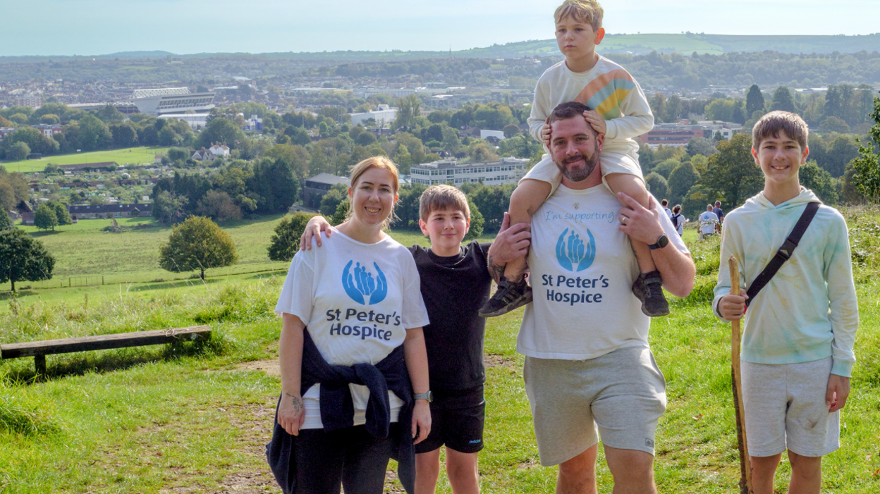 A family with three boys -one of whom is sitting on his father's shoulders - stand on a hillside with Bristol in the background. The parents are wearing white t-shirts with the St Peter's Hospice logo on. 