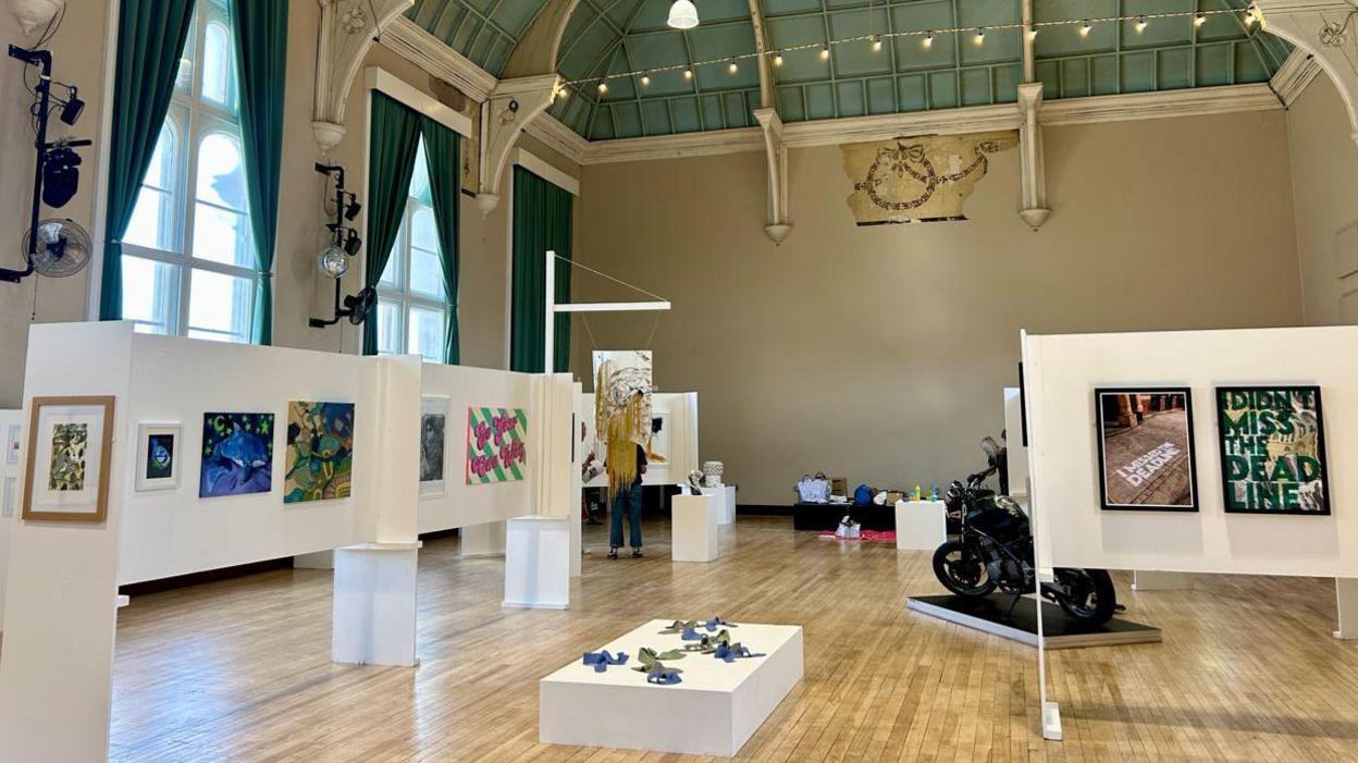 A selection of artwork, including paintings and photographs, are on display as part of an exhibition. A motorbike can also been seen on display. 