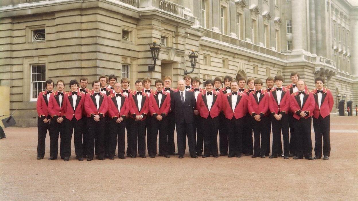 A group of musicians wearing red jackets, bow ties and black trousers, with one wearing a black suit and a tie, standing outside Buckingham Palace.