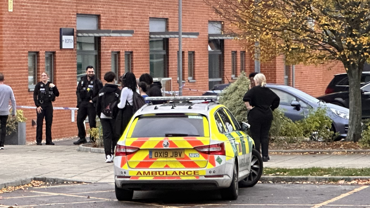 An ambulance parked outside the college with two police officers and a group of students and staff outside the college, in front of a cordon.