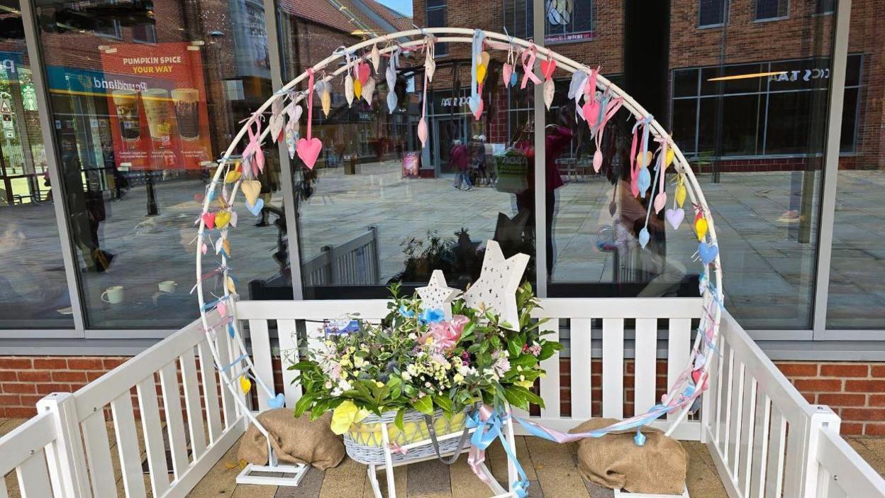 A circular white display with colourful ribbons and hearts with a cot with green leaves, flowers and stars elevated on a white platform