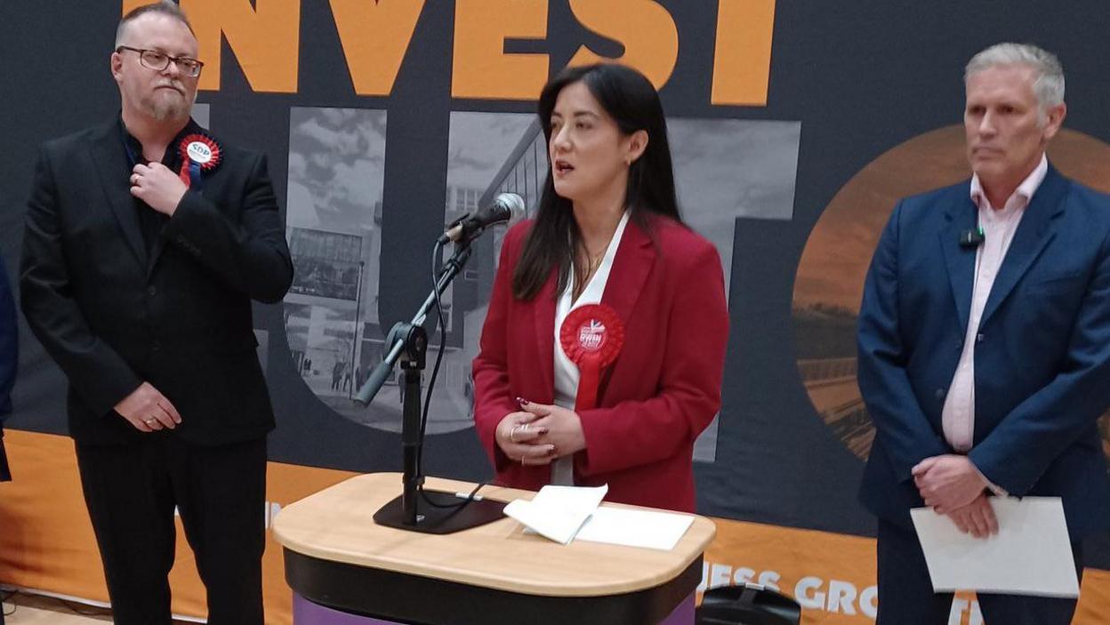 Sarah Owen with her hands clasped together stands at a podium and speaks into a microphone. She is wearing a red jacket with a red rosette attached to its lapel, two men stand either side of her.