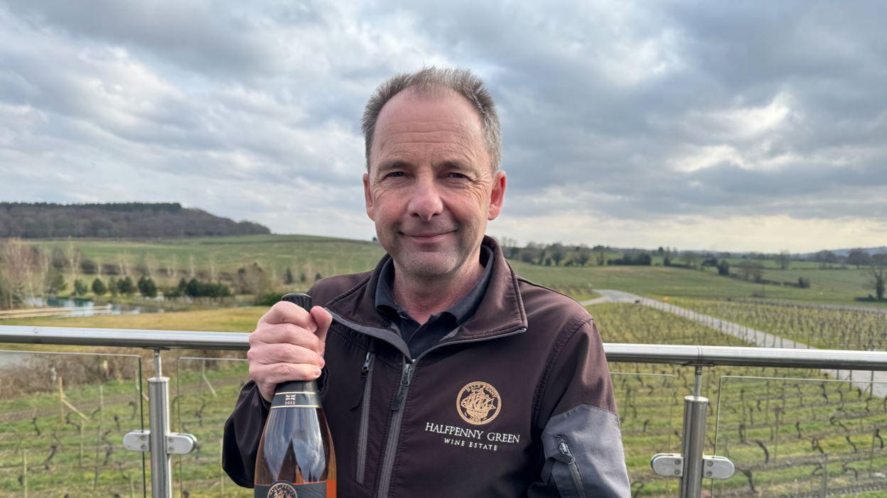 A man stands in a vineyard holding a bottle of wine up to the camera.