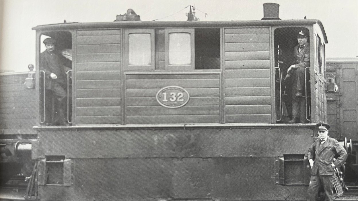 A black and white photograph of a tram engine which is in the middle. Two people are standing in the doorways of the engine either side and a person is stood on the ground on the right.