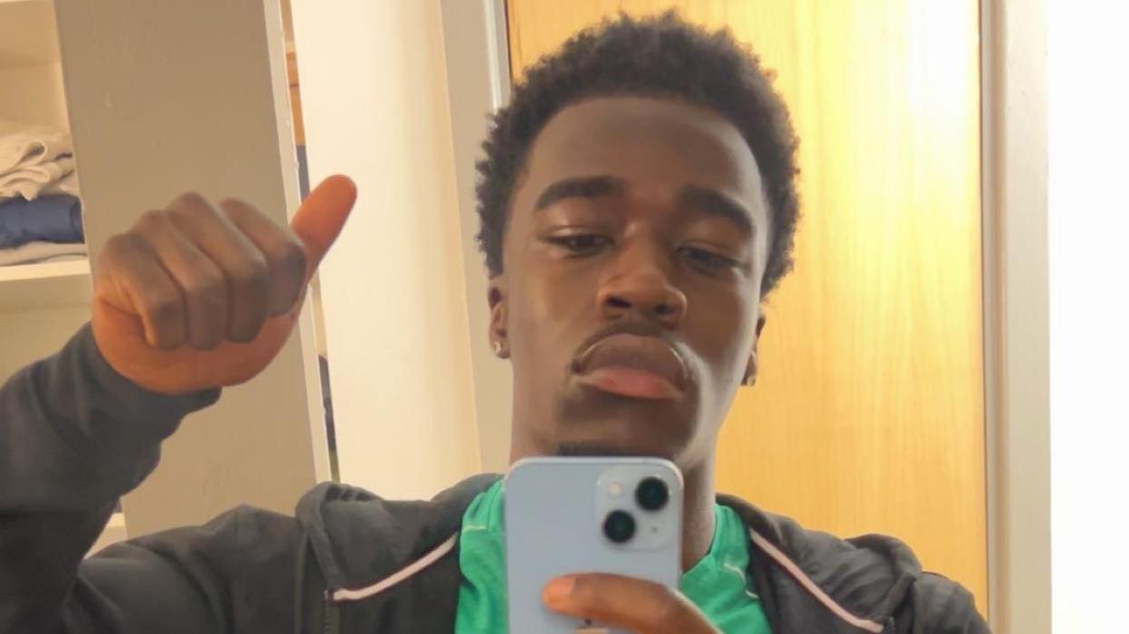 Joshua Adiefeh sticks his thumb while holding a phone to take a selfie. He is wearing a green T-shirt and grey hoodie
