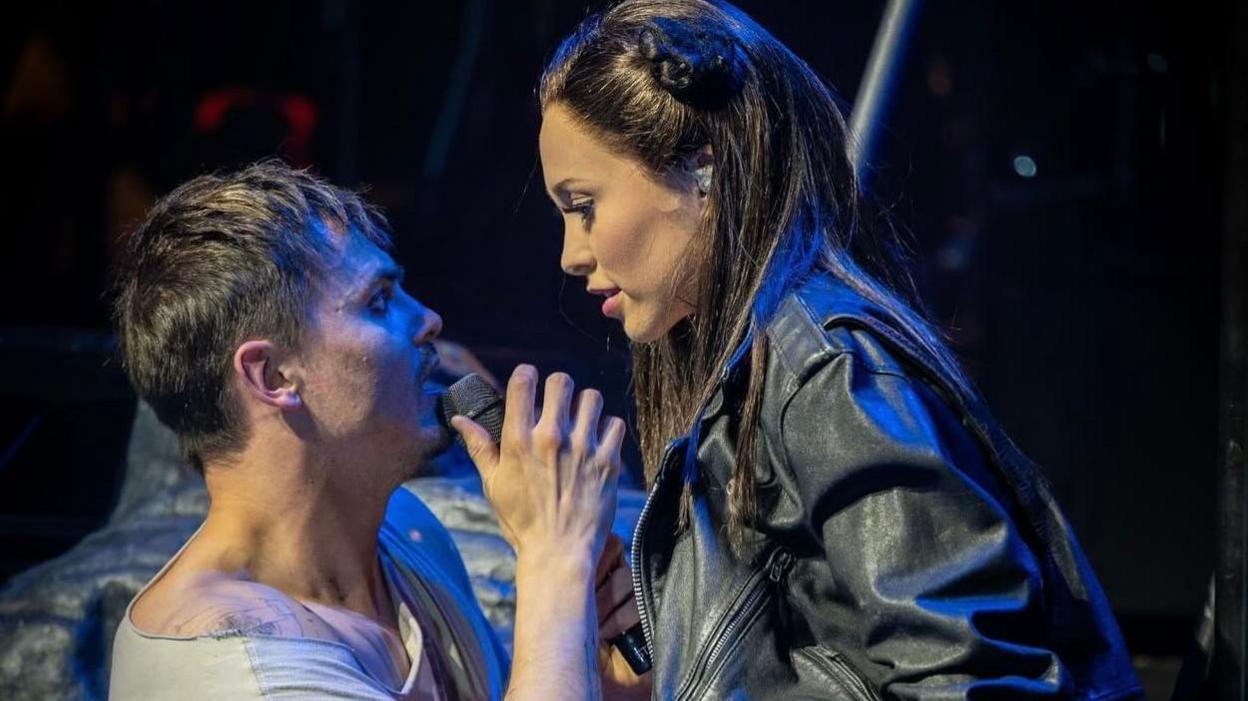 An actress with dark hair wears a leather jack and fishnet tights on stage. She looks down at a male actor with brown hair wearing a white top, who has tattoos on his arm. The stage behind them is dark.