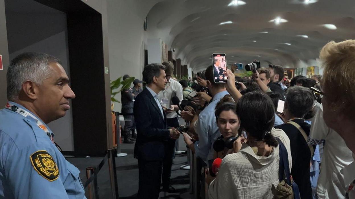 Mr Miliband stands wearing a suit stands behind a security line with a crowd of journalists.
