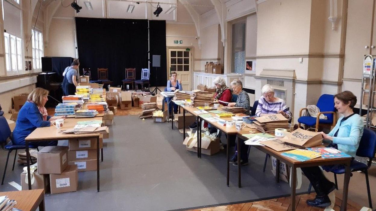 Let's Talk Reading volunteers sit in a large hall behind tables that are lined up in the room facing each other. The tables are covered with books and bags. The volunteers, who are all women, are packing the bags with the books. The hall has cream walls and a large black screen at the back.