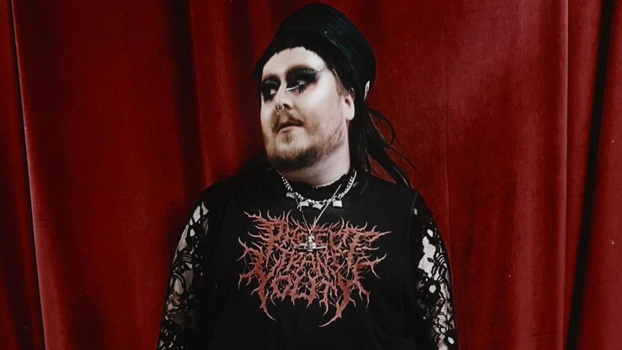 A person with gothic make-up, several chains around their neck and a top with horror-style red lettering stands in front of a blood-red curtain, looking off to the side.