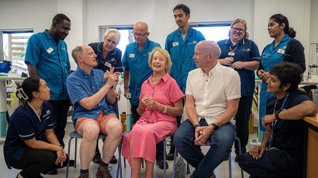 A group of medical professionals sit on chairs in a blood donation ward, with three members of the public sitting together on the front row