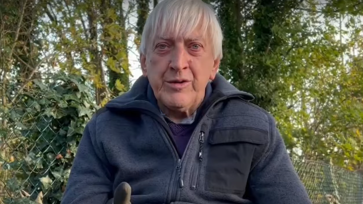 Geoff, a man with grey hair. He is wearing a navy jacket and there are several green trees and shrubs behind him.