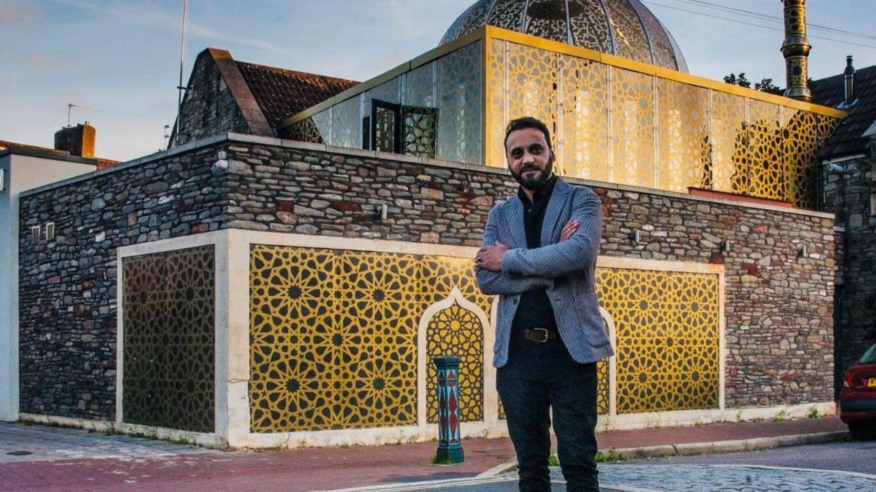 Abdul Malik wearing dark trousers and a grey blazer standing with his arms folded in front of a mosque, with gold gilded patterns and bricks