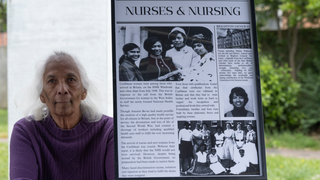 Shirley Williams sits next to a framed image titled "nurses & nursing," describing how migrants from the Caribbean and across the Commonwealth who came to Britain on the HMT Empire Windrush contributed to the National Health Service (NHS) post-World War II. It features black and white images of Shirley as a young woman.