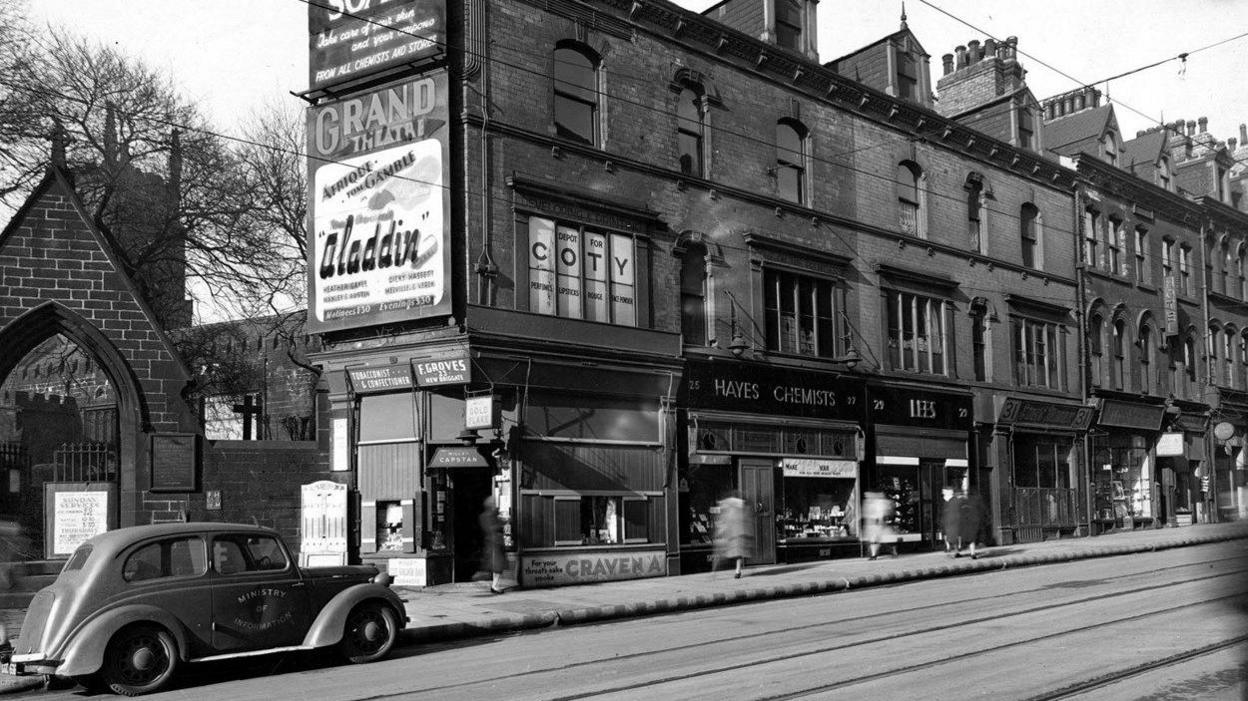 A black and white image of New Briggate, including a row of shops and a churchyard gate, with a 1940s-style car parked to the left and tramlines running in front of the shops.