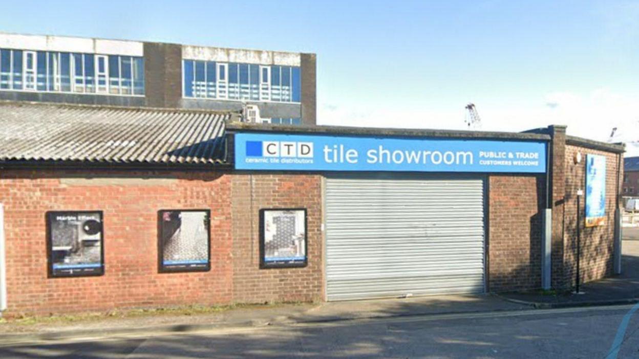 A single brick building with a blue banner over a shuttered garage saying tile showroom. It looks very run down 