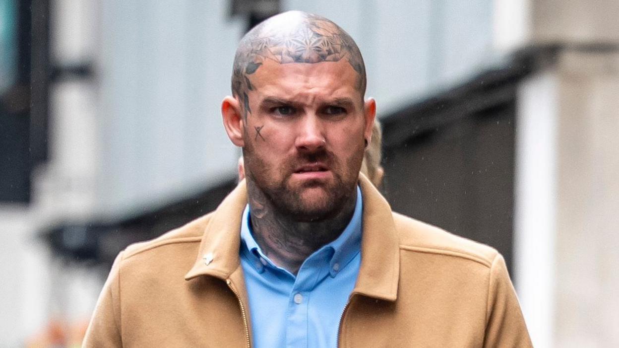 Tony Bartlett walks towards Bristol Crown Court. He is a large man with a shaved head and several tattoos on his neck and head, but not on his face itself. He is wearing a light blue shirt under a light brown jacket.