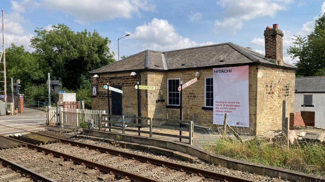 A small stone brick building sits behind railway lines on a sunny day. There's a railway crossing to the right with trees.