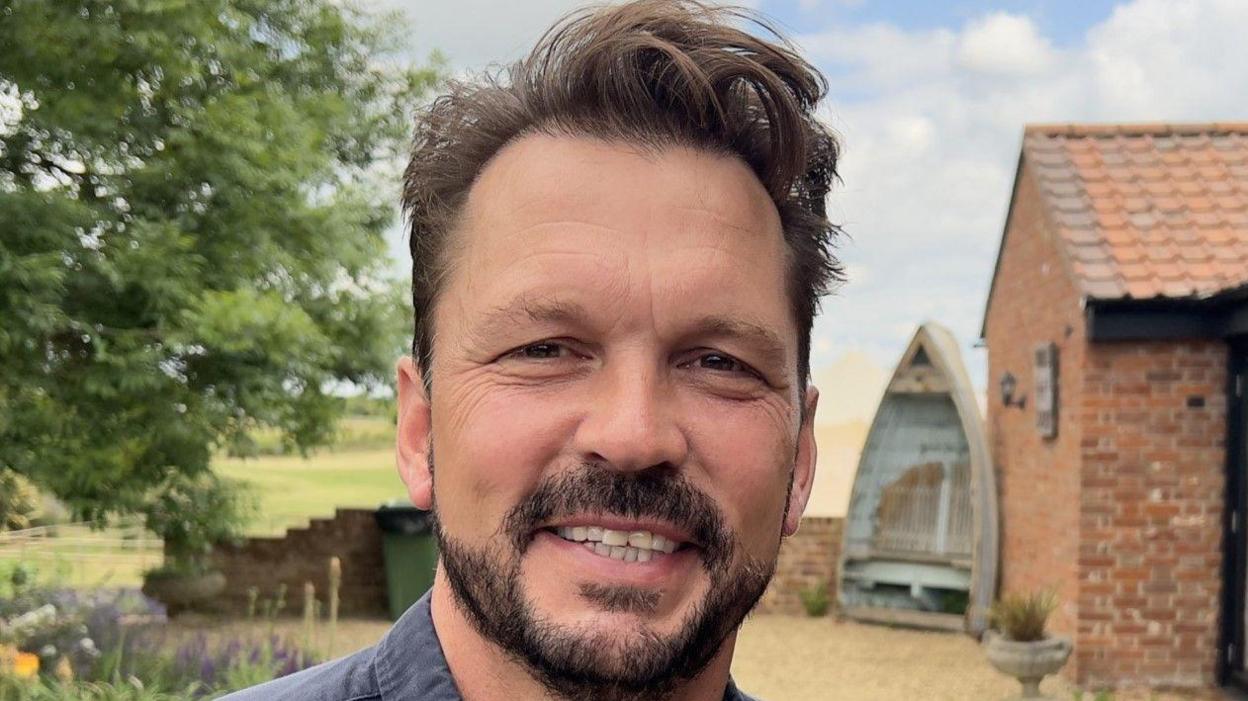 Jimmy Doherty smiles at the camera. He has short brown hair and a brown beard. He wears a blue shirt.