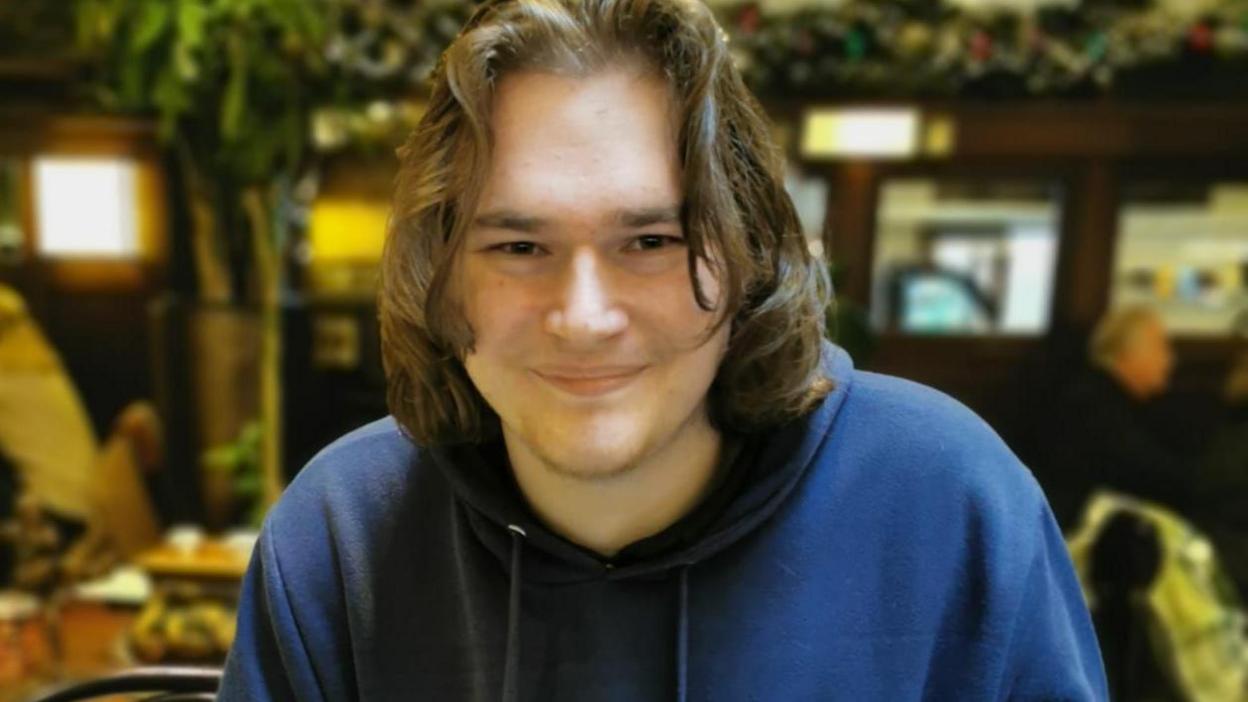 A man in his early 20s, who has brown eyes,  long brown hair and is clean shaven, smiling into the camera. He is sitting on a chair in a cafe and is wearing a blue hoody. 