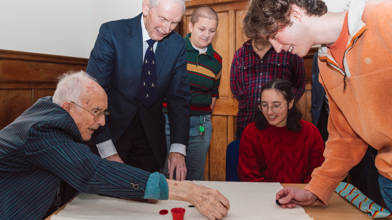 Bill Steen, far left, with Peter Downes, playing tiddlywinks with current Cambridge University staff and students. They are sat around a wooden table, with a white mat atop, trying to get the circular pieces into a small red cup. 