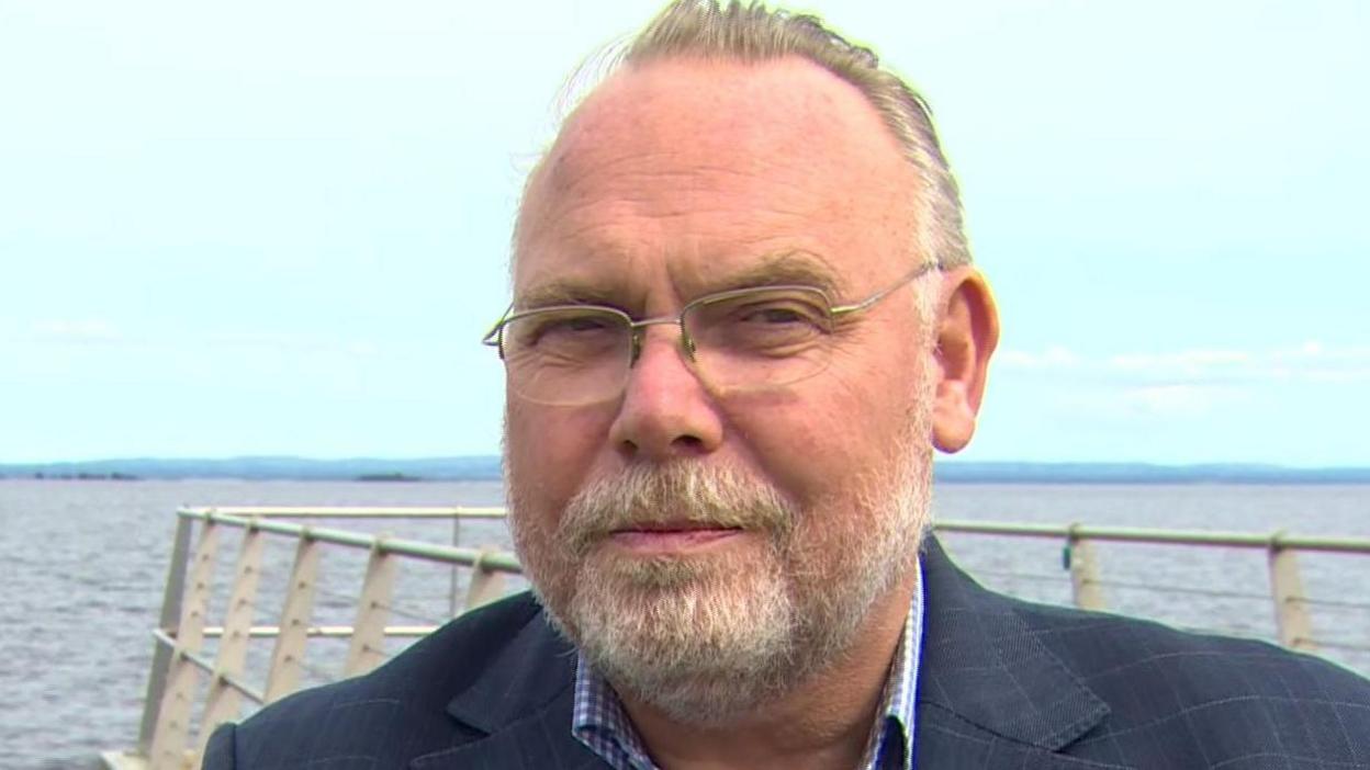 Gerry wear glasses and has a beard. He's wearing a suit and is standing with the lough behind him.