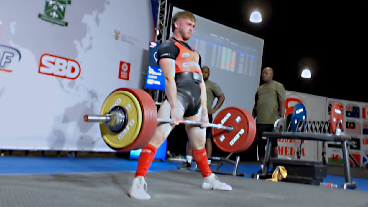 Lucas Williams completing a deadlift, standing upright with a barbell in both hands, weighted on both sides with several weight plates. He is wearing a red Cymru vest, a black T-shirt and shorts, with a lifting belt, and red long socks and white training shoes. Behind him stand two men at the side of stage, with competition logos on a screen behind him