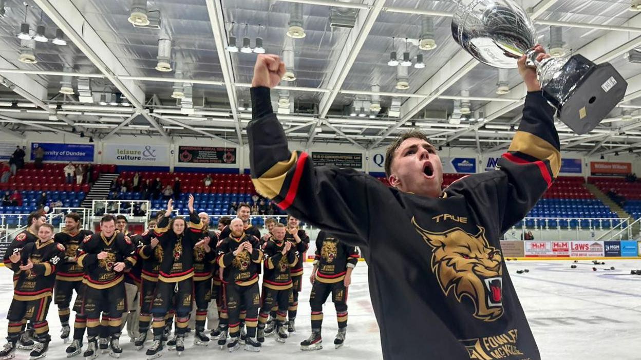 Gary Kelly standing with his Aberdeen Lynx Ice Hockey teamates on an ice rink, holds aloft a trophy in triumph.