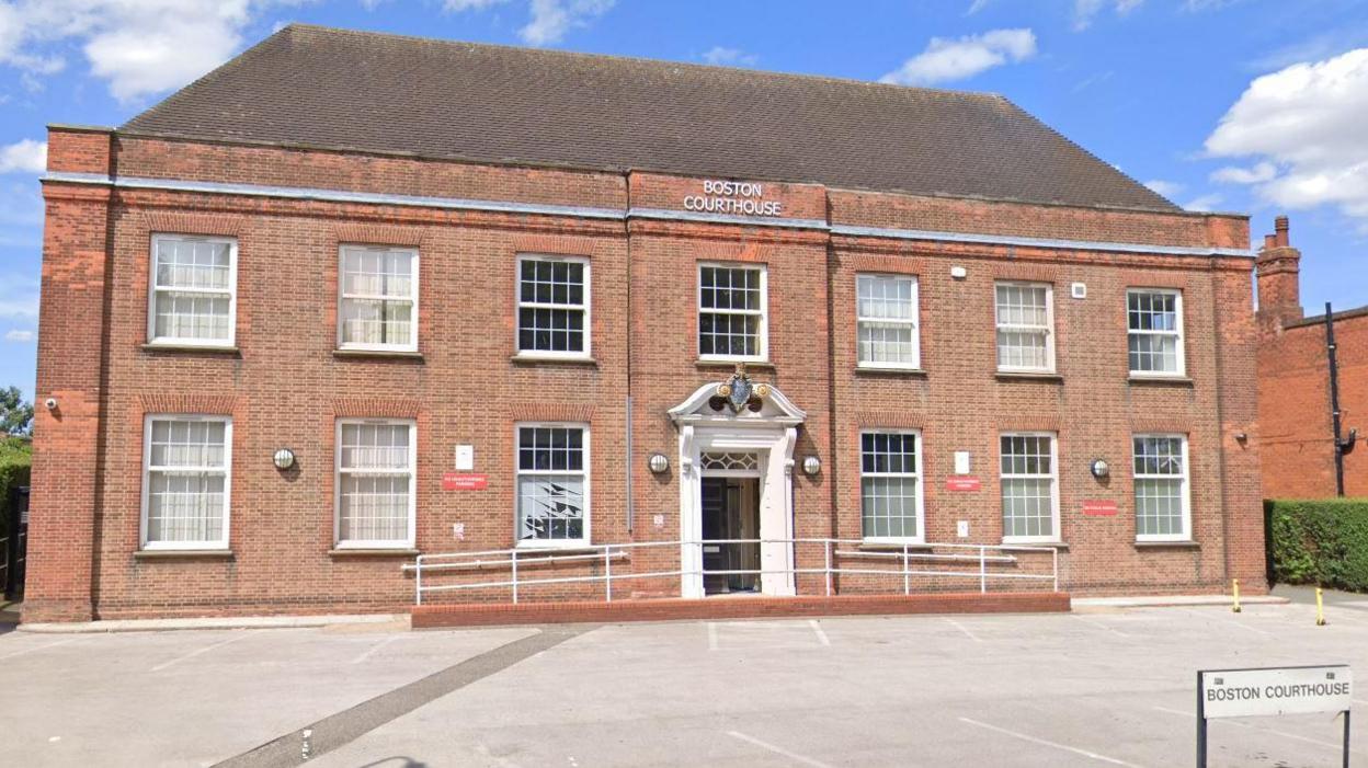 A large two-storey redbrick Georgian-style building. On the front below the roof a signage reads 'BOSTON COURTHOUSE'. Underneath is a window and below that is the front porch which is white and decorative - on the top arch of the door is the court crest logo. There are empty parking bays in front of the building.