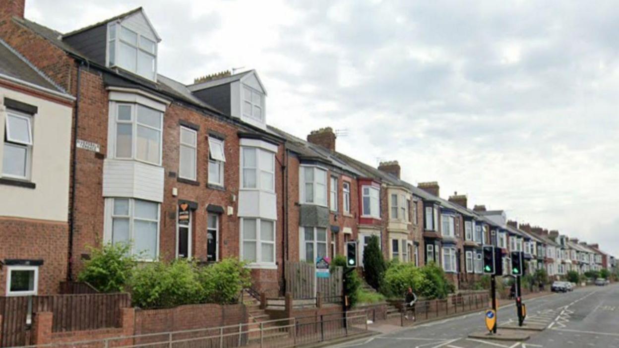 A row of terraced housing is on the left hand side of the road. There is a pedestrian crossing with traffic lights in the centre of the road.
