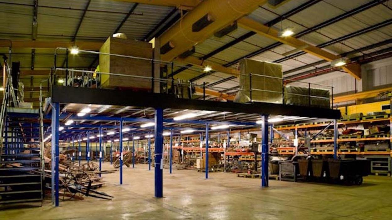 A large warehouse with large objects, you can see boxes stacked on shelves, there are blue pillars throughout the building.