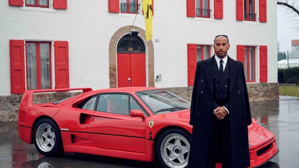 Lewis Hamilton stood alongside a Ferrari sports car while wearing a black suit and black overcoat