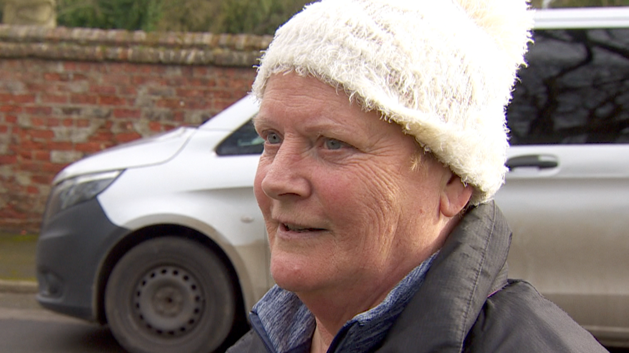 Sue has blue eyes and is wearing a white woolly hat on her head, a grey coat over a blue top.