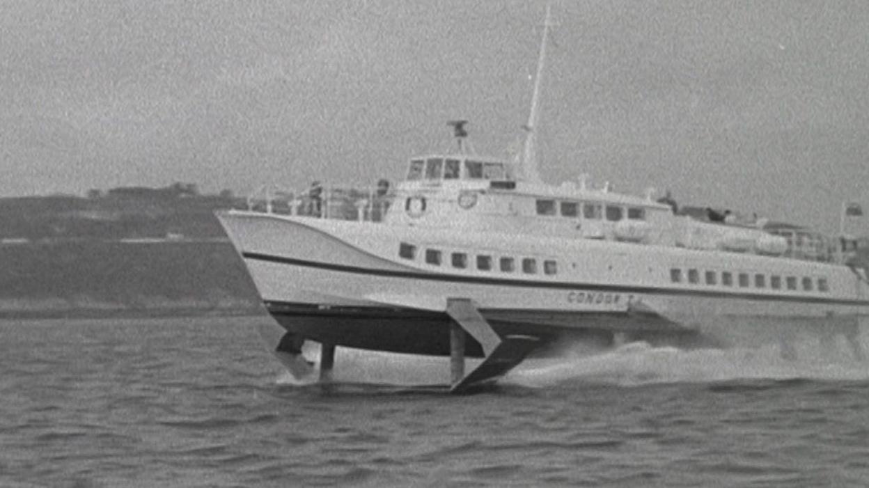 Black and white image of a white vessel on the waters, with land visible in the background.